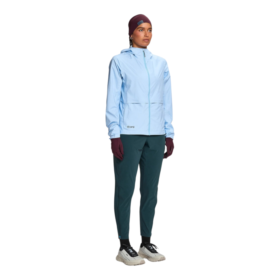 Up & Running model wearing a light blue Janji Rainrunner Pack Jacket with a hood, dark teal trousers, and beanie.